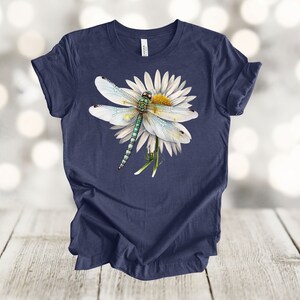 May include: Navy blue t-shirt featuring a detailed illustration of a dragonfly resting on a white daisy. The dragonfly has a green and blue body with delicate, translucent wings. The daisy has a yellow center and white petals. The shirt is made of soft material.