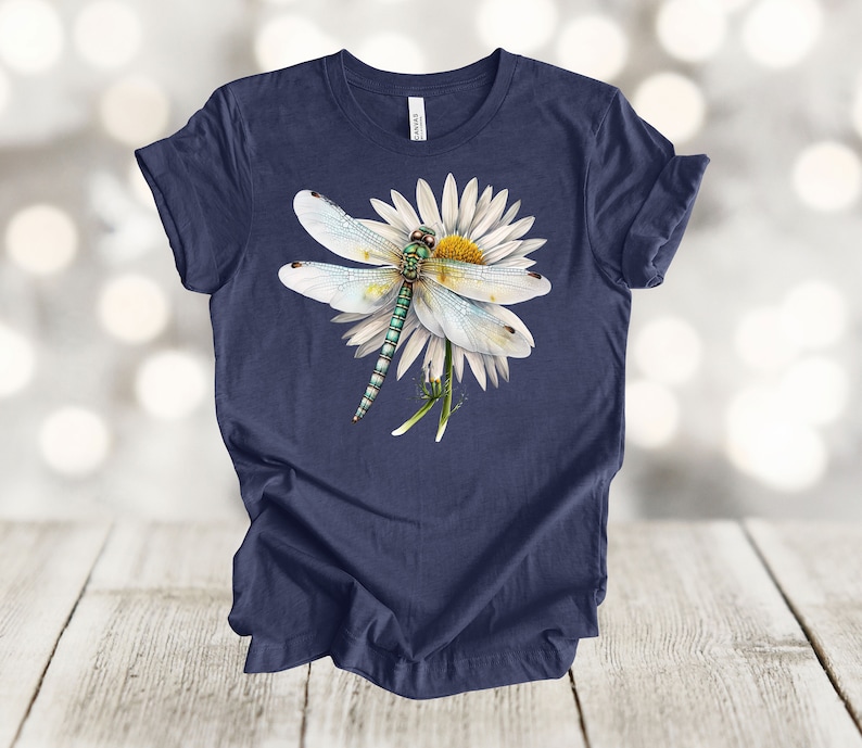 May include: Navy blue t-shirt featuring a detailed illustration of a dragonfly resting on a white daisy. The dragonfly has a green and blue body with delicate, translucent wings. The daisy has a yellow center and white petals. The shirt is made of soft material.