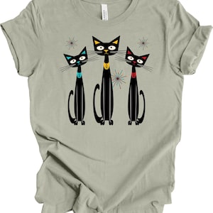 May include: A light gray t-shirt featuring a retro design of three black cats with colorful collars and starburst accents. The cats have yellow, blue, and red collars. The shirt is made of a soft material.