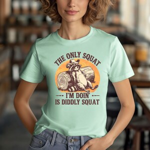 May include: A light green t-shirt with a graphic of a raccoon wearing a cowboy hat and sitting on a log. The text on the shirt reads "The only squat I'm doin' is diddly squat".