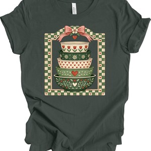 May include: Dark green t-shirt featuring a graphic of stacked decorative bowls. The bowls are in shades of green and white, adorned with floral and heart patterns. A pink bow and a checkered border frame the design.