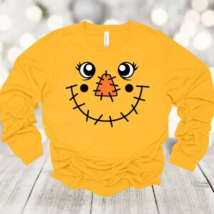 May include: Mustard yellow long-sleeve shirt featuring a smiling scarecrow face graphic. The graphic includes black stitched details, large eyes, and a stitched nose. The shirt is made of a soft material.