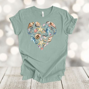May include: A light green t-shirt with a heart-shaped design made up of seashells, starfish, seahorses, and anchors. The design is in shades of blue, brown, pink, and yellow.