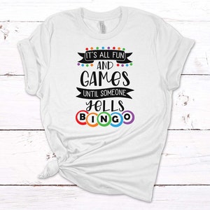 May include: White t-shirt with a black graphic that says "It's all fun and games until someone yells bingo". The graphic includes colorful bingo balls.