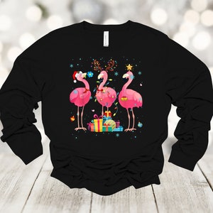 May include: Black long-sleeved shirt with a Christmas-themed design featuring three pink flamingos wearing Santa hats and reindeer antlers, surrounded by Christmas lights and presents.