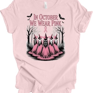 May include: Light pink t-shirt with the text "In October We Wear Pink" and a graphic of five witches in pink robes and hats. The design includes bats and a pink ribbon, set against a forest backdrop.