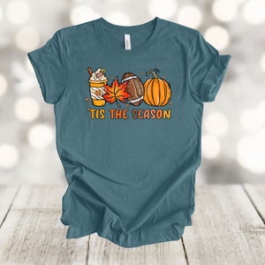May include: Teal blue t-shirt with a graphic of a pumpkin, football, fall leaves, and a coffee drink. The text "'Tis the season" is printed below the graphic.