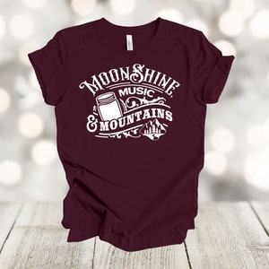 May include: Burgundy t-shirt with a white graphic design that says "Moonshine, Music & Mountains". The graphic includes a mason jar and a mountain scene.