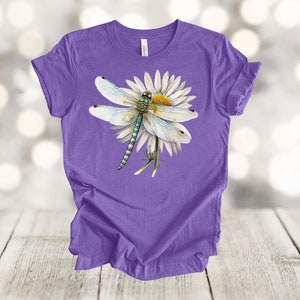 May include: A purple t-shirt featuring a detailed illustration of a dragonfly perched on a white daisy with a yellow center. The dragonfly has light blue wings and a green and blue body. The shirt is made of a soft material.