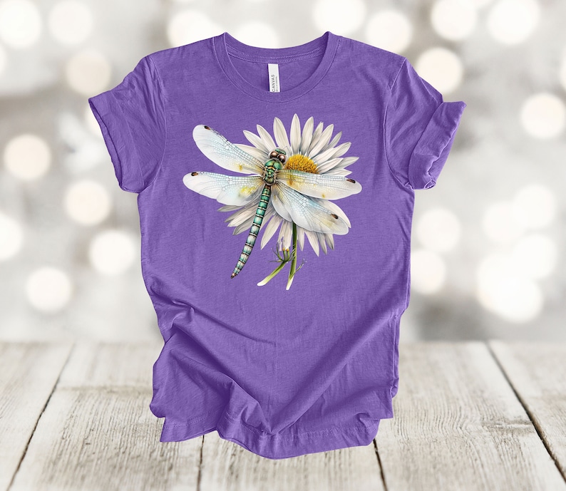 May include: A purple t-shirt featuring a detailed illustration of a dragonfly perched on a white daisy with a yellow center. The dragonfly has light blue wings and a green and blue body. The shirt is made of a soft material.