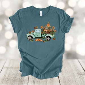 May include: Teal t-shirt featuring a vintage teal pickup truck filled with pumpkins, sunflowers, and a scarecrow. The truck is decorated with fall foliage. A charming autumn-themed graphic for seasonal wear.
