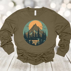 May include: A long-sleeved olive green t-shirt with a graphic print of a mountain range with a sunset behind it. The graphic is in a circle with a blue and green background.