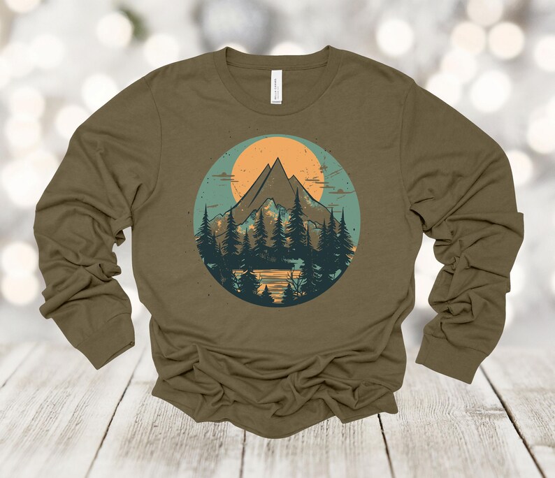 May include: A long-sleeved olive green t-shirt with a graphic print of a mountain range with a sunset behind it. The graphic is in a circle with a blue and green background.