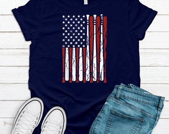 May include: Navy blue t-shirt featuring a distressed American flag design made of baseball bats. The flag design incorporates the stars and stripes, with the stripes represented by red and white bats. The shirt is paired with blue jeans and white sneakers.