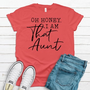 May include: A coral colored t-shirt with black text that reads "Oh honey, I am that aunt".