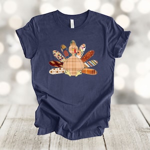 May include: A navy blue t-shirt with a patchwork turkey design. The turkey is brown and white with a red wattle and a yellow beak. The feathers are a variety of colors and patterns, including floral, plaid, and polka dots.