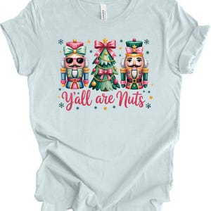 May include: Light blue t-shirt with a colorful graphic featuring two nutcrackers and a Christmas tree. The text "Y'all are Nuts" is written in pink and red. The shirt is made of a soft material.