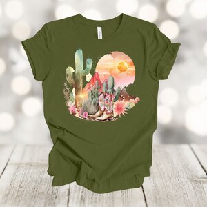 Camiseta gráfica Atardecer en el desierto: Camiseta estilo vaquero con estampado de cactus y botas. Olive