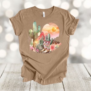 Camiseta gráfica Atardecer en el desierto: Camiseta estilo vaquero con estampado de cactus y botas. Heather Tan