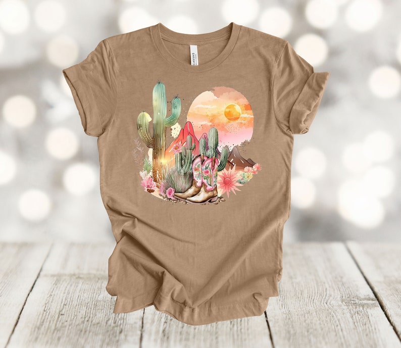 Camiseta gráfica Atardecer en el desierto: Camiseta estilo vaquero con estampado de cactus y botas. Heather Tan