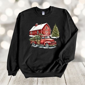 May include: A black sweatshirt featuring a red barn, a red truck with a Christmas tree in the back, and a green Christmas tree in the background. The truck is driving through a snowy scene.