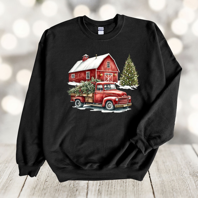 May include: A black sweatshirt featuring a red barn, a red truck with a Christmas tree in the back, and a green Christmas tree in the background. The truck is driving through a snowy scene.