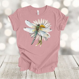 May include: A dusty rose t-shirt featuring a detailed illustration of a dragonfly with white wings and a green body, perched on a white daisy with a yellow center. The design is centered on the shirt.