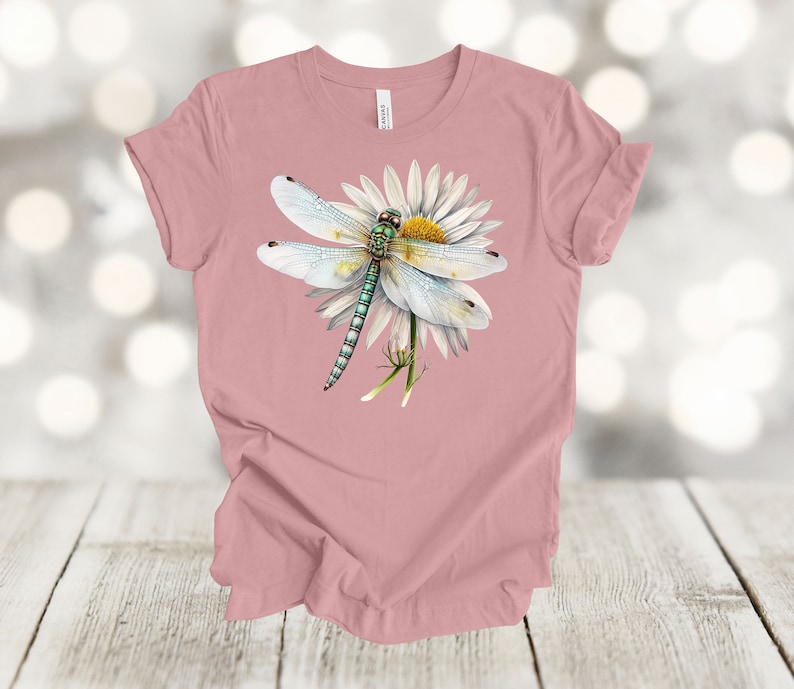 May include: A dusty rose t-shirt featuring a detailed illustration of a dragonfly with white wings and a green body, perched on a white daisy with a yellow center. The design is centered on the shirt.