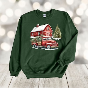 May include: A green sweatshirt featuring a red truck carrying a Christmas tree in front of a red barn with a snowy roof. The truck is driving through a snowy landscape with a Christmas tree in the background.