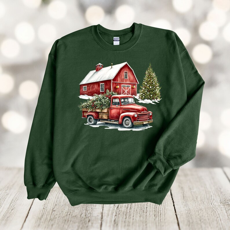 May include: A green sweatshirt featuring a red truck carrying a Christmas tree in front of a red barn with a snowy roof. The truck is driving through a snowy landscape with a Christmas tree in the background.