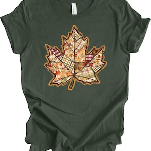 May include: Dark green t-shirt featuring a large, patchwork maple leaf design. The leaf is composed of various autumn-themed patterns in shades of red, orange, yellow, and green, with a brown outline. A casual, fall-themed apparel item.