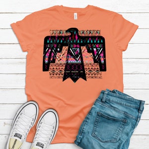May include: A peach colored t-shirt with a black silhouette of an eagle with a geometric pattern in pink, blue, and turquoise. The eagle is centered on the shirt. The shirt is laid flat on a white wooden surface with a pair of white canvas shoes and a pair of blue jeans.