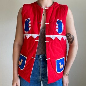 May include: A red quilted vest with blue and white applique designs. The vest has a button closure and two pockets.