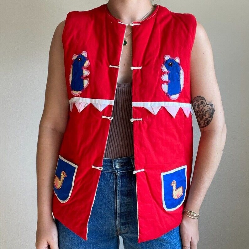 May include: A red quilted vest with blue and white applique designs. The vest has a button closure and two pockets.