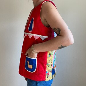 May include: A red quilted vest with a patchwork design. The vest has a blue pocket with a yellow bird applique and a white trim. The vest is sleeveless and has a V-neck.