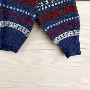 May include: Blue, red, white, and gray patterned sweater with a knitted texture. The sweater has a long sleeve and a ribbed cuff.