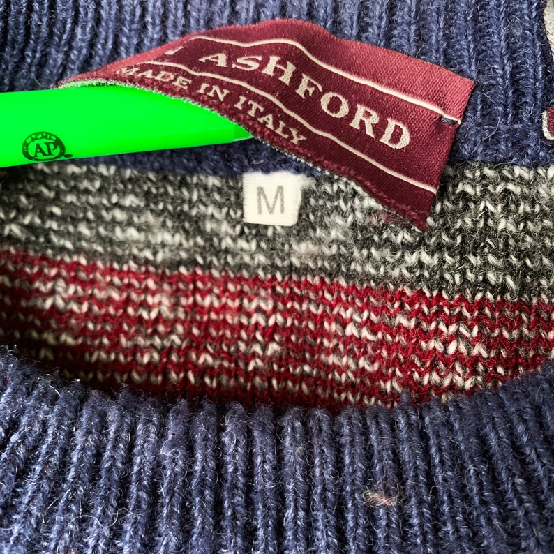 May include: A blue, red, and gray striped sweater with a burgundy label that reads "Ashford Made in Italy" and a white tag with the letter "M" on it.