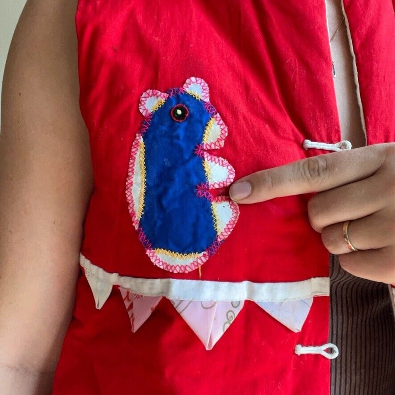 May include: Red vest with a blue and white embroidered animal applique. The vest has a white trim and pink and white scalloped edges.