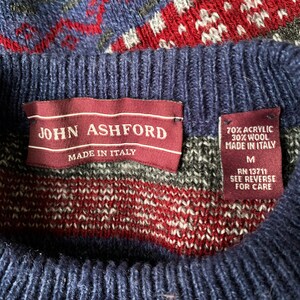May include: A navy blue sweater with a red, white, and gray geometric pattern. The sweater has a label that reads "JOHN ASHFORD MADE IN ITALY" and a care label that reads "70% ACRYLIC 30% WOOL MADE IN ITALY M RN 13711 SEE REVERSE FOR CARE".