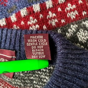 May include: A close-up of a burgundy care label attached to a blue and red knitted sweater. The label reads "Machine Wash Cold Gentle Cycle Do Not Bleach Tumble Dry Low".