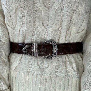 Puede incluir: Un cintur&oacute;n de cuero marr&oacute;n con una hebilla plateada adornada. El cintur&oacute;n se lleva sobre un su&eacute;ter de punto de cable de color crema.