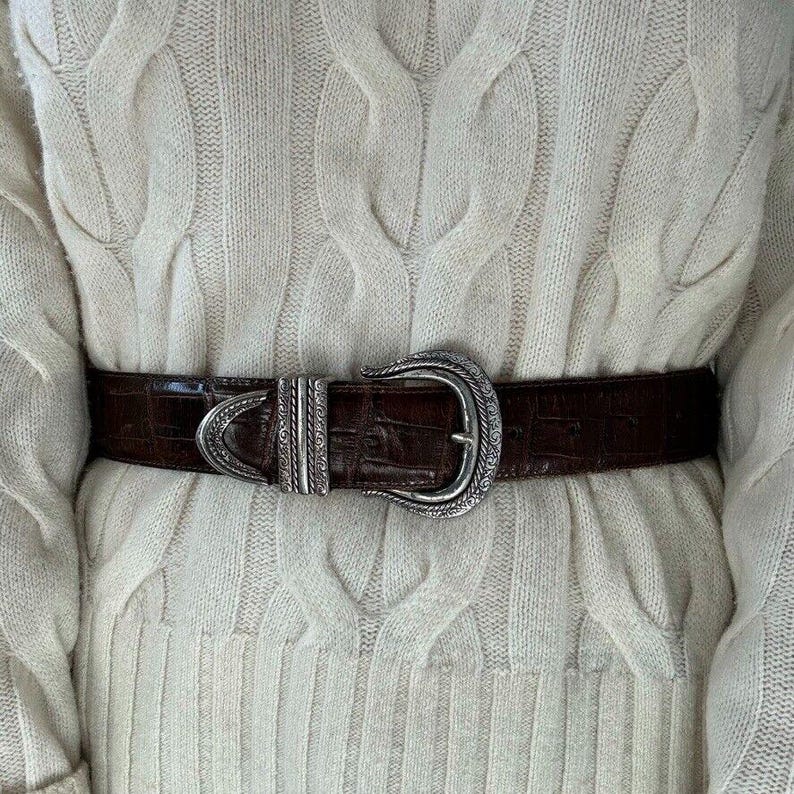 Puede incluir: Un cintur&oacute;n de cuero marr&oacute;n con una hebilla plateada adornada. El cintur&oacute;n se lleva sobre un su&eacute;ter de punto de cable de color crema.
