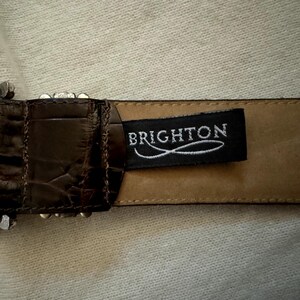 Puede incluir: Un cintur&oacute;n de cuero marr&oacute;n con una hebilla plateada y una etiqueta de cuero negro que dice "BRIGHTON".