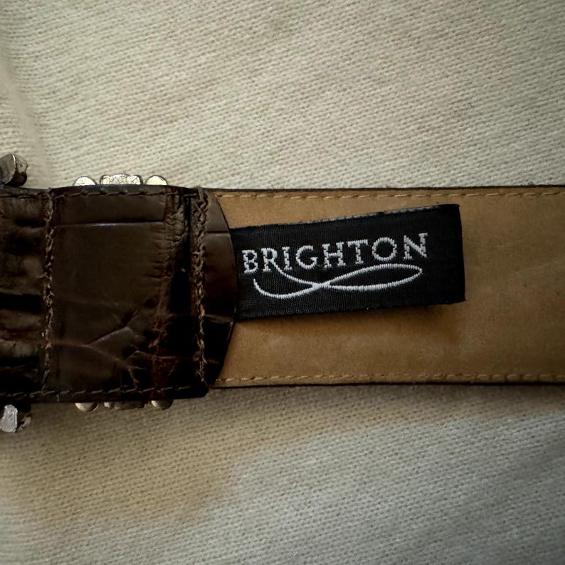 Puede incluir: Un cintur&oacute;n de cuero marr&oacute;n con una hebilla plateada y una etiqueta de cuero negro que dice "BRIGHTON".