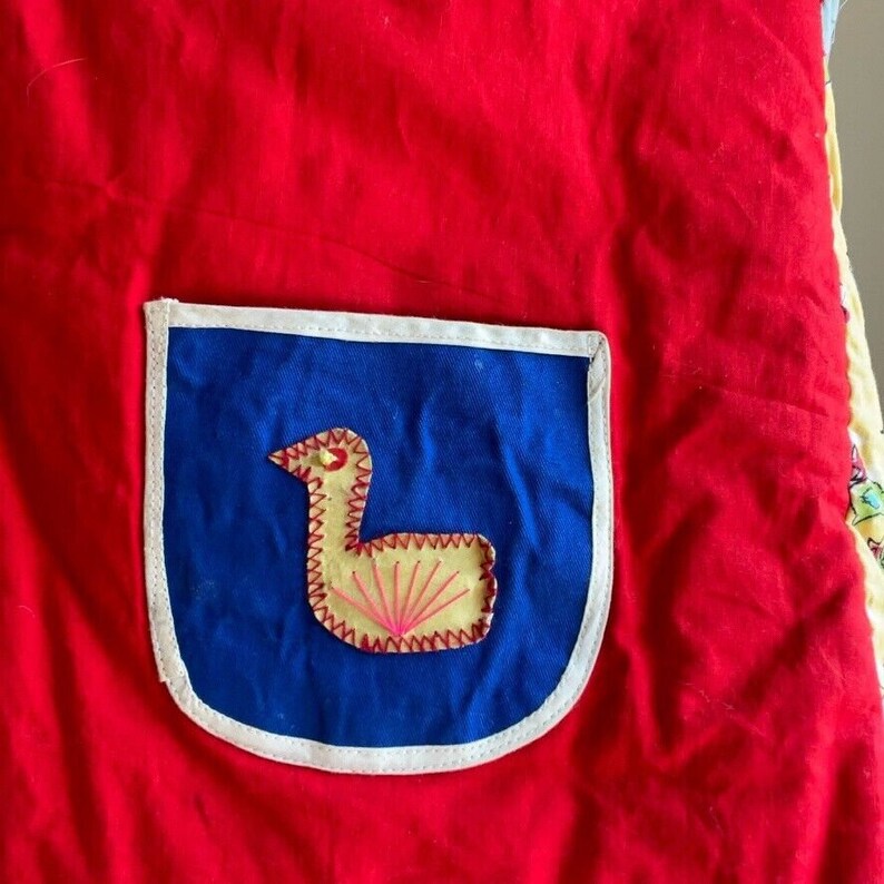 May include: A red fabric with a blue and white pocket. The pocket has a yellow embroidered bird with pink accents.