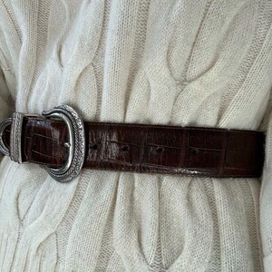 Puede incluir: Un cintur&oacute;n de cuero marr&oacute;n con una hebilla plateada adornada. El cintur&oacute;n se lleva sobre un su&eacute;ter de punto de cable blanco.