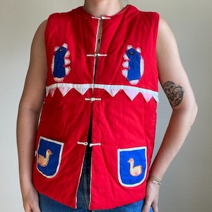 May include: A red quilted vest with blue and white applique designs. The vest has a mandarin collar and a button closure. The vest has two pockets on the front with blue and white applique designs.
