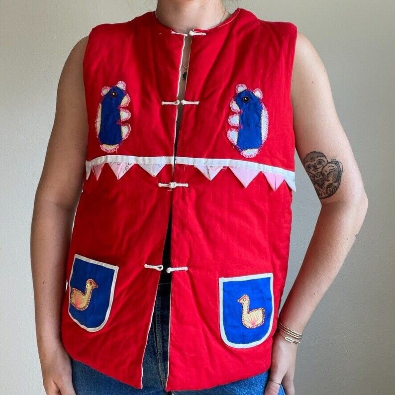May include: A red quilted vest with blue and white applique designs. The vest has a mandarin collar and a button closure. The vest has two pockets on the front with blue and white applique designs.