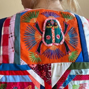 May include: A colorful patchwork vest with a large embroidered frog design on the back. The frog is green with pink and black accents and is surrounded by colorful embroidery.