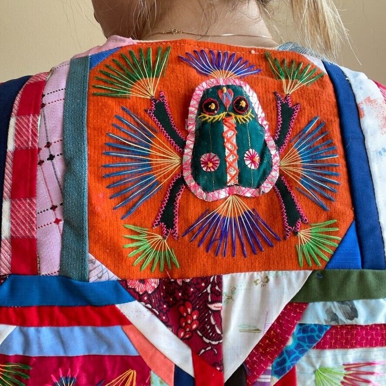 May include: A colorful patchwork vest with a large embroidered frog design on the back. The frog is green with pink and black accents and is surrounded by colorful embroidery.